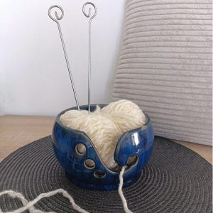 Könnte beinhalten: Eine tiefblaue Keramik-Garnschale mit glänzender Oberfläche, die einen spiralförmigen Ausschnitt und mehrere kreisförmige Löcher an der Seite aufweist. In der Schale liegt ein cremefarbenes Garnknäuel mit zwei langen, dünnen Stricknadeln aus Metall.