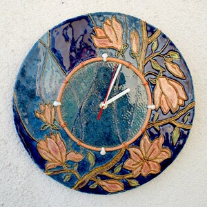 May include: A blue ceramic clock with a white face and gold accents. The clock is decorated with pink magnolia flowers and green leaves.