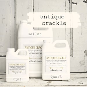 May include: Three white plastic bottles of antique crackle paint in pint, quart, and gallon sizes. The bottles are labelled with the product name and the size. The bottles are sitting on a white wooden surface.