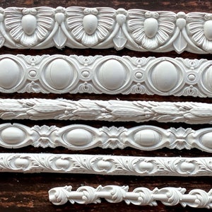 May include: Six white decorative molding strips with intricate designs, including floral, geometric, and oval patterns. The strips are arranged horizontally on a brown wooden surface.