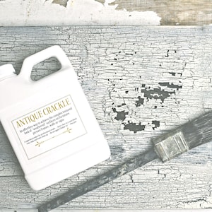 May include: A white plastic bottle with a gold label that reads "Antique Crackle". The label describes the product as an effortless way to achieve a weathered look on furniture, picture frames, walls, or signs. A paintbrush lies on a white, cracked surface.