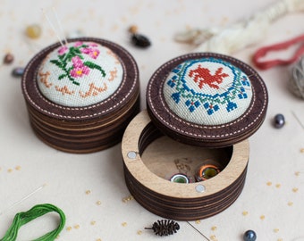 May include: Two round wooden boxes with lids, each featuring a different cross-stitch design. One lid has a floral design with pink flowers, the other has a red bird design. The boxes are open, revealing a compartment for storing sewing supplies.