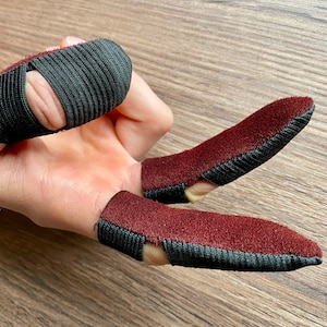 May include: A pair of fingertip protectors, one black and one burgundy, with a black elastic band around the base of each. The protectors are worn on the index and middle fingers.