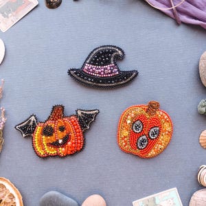 Puede incluir: Tres broches de Halloween con cuentas: un sombrero de bruja, una calabaza con alas de murciélago y una calabaza con una cara. El sombrero de bruja es negro con una banda rosa. Las calabazas son naranjas y rojas con detalles negros.
