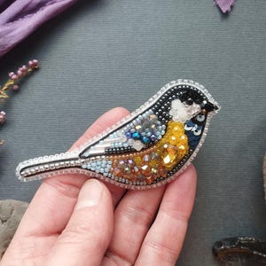 A hand is holding a decorative bird brooch made of colorful beads and sequins, with a purple fabric and some dried flowers in the background.