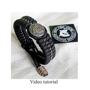 May include: Black paracord bracelet with a gold-toned metal charm featuring a triangular symbol. The bracelet has a brown and black braided design. The charm has the text "Knot Findz Viking Aggressive Bracelet".