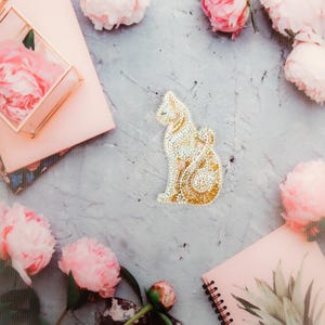 Könnte beinhalten: Eine weiße Katzenbrosche mit goldenen Akzenten liegt auf einer grauen Oberfläche, umgeben von rosa Pfingstrosen und geöffneten Notizbüchern.