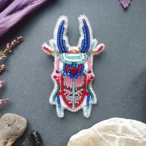 a colorful, intricately designed brooch or pin with a detailed, beaded pattern. The brooch is placed on a gray surface, surrounded by various objects such as rocks, leaves, and flowers.