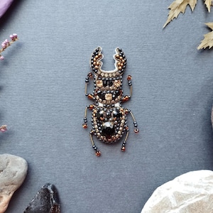 a detailed embroidered or beaded insect, possibly a beetle, set against a grey background with various natural elements such as leaves, rocks, and flowers.