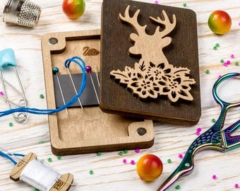 Op de afbeelding: Een houten doos met een magnetische sluiting, met een lasergesneden ontwerp van een hert met bloemmotieven. De doos is open en laat een magnetische naaldhouder zien met blauwe draad en spelden. De doos is omringd door kleurrijke kralen en een paar regenboogkleurige scharen.
