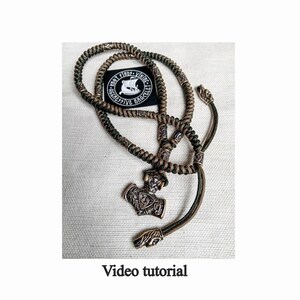 May include: A green and brown braided cord necklace with a metal pendant shaped like a hammer. The pendant has intricate details and is attached to the cord with a metal ring. The necklace is on a beige fabric background. The text "Knot Finds Viking Bracelet" is visible on a black sticker.