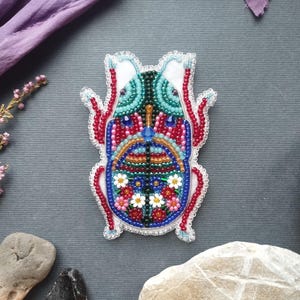 a colorful, intricately beaded object resembling a bug or insect, surrounded by various natural elements such as flowers, rocks, and leaves.