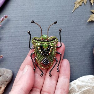 A hand holding a green and black beaded insect brooch against a gray background with various natural elements such as leaves and rocks.