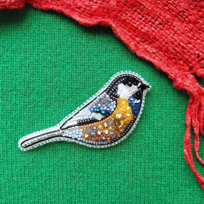 A colorful, hand-made bird brooch with a beaded and embroidered design rests on a green fabric surface, with a red knitted item partially visible in the background.