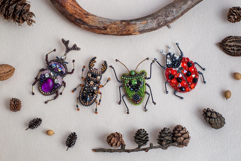 four colorful, intricately designed insects, including a ladybug, a beetle, and two other bugs, arranged on a white background. The insects are surrounded by various natural elements, such as pine cones, acorns, and a tree branch.