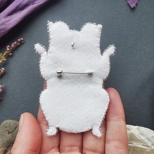 A hand holding a white felt animal pin against a dark background with some purple flowers and a brown object.