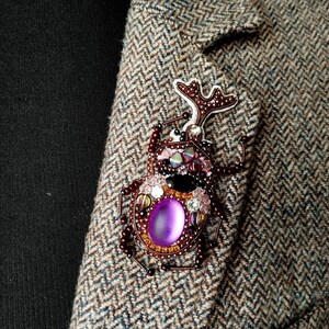 A close-up image of a decorative insect brooch with a purple gemstone, placed on a tweed jacket.