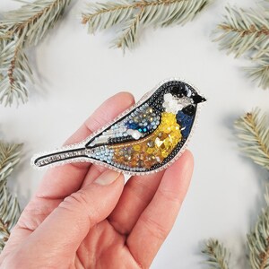 a hand holding a bird brooch with a pine branch in the background