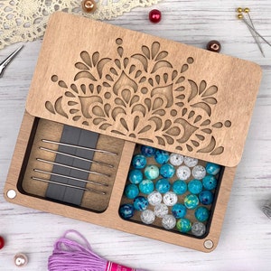 May include: A wooden box with a cutout design on the lid. The box has two compartments, one for needles and one for beads. The beads are blue, white, and green.
