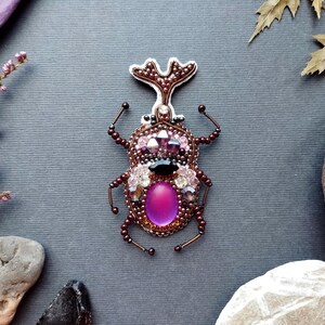 a detailed embroidered insect brooch with a purple center and various embellishments, including gemstones and beads, set against a gray background.