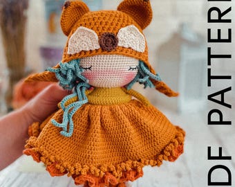 May include: Crocheted doll with a fox hat, orange dress, and blue hair. The doll is holding a small, crocheted flower. The doll is standing on a wooden surface. PDF PATTERN.