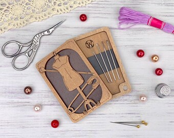 Op de afbeelding: Een houten naaiset met een uitsnijding van een paspop, een veiligheidsspeld en een naald. De set bevat een compartiment voor naalden en garen. De set ligt op een wit houten oppervlak met roze garen, een vingerhoed en kralen.