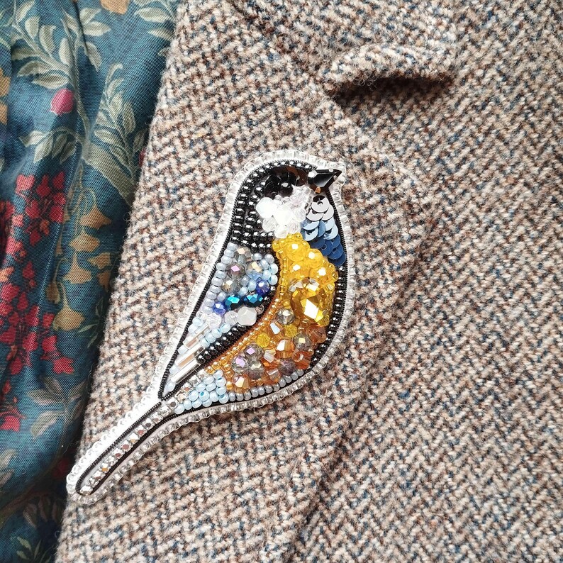 A close-up image of a decorative bird pin or brooch, featuring a detailed and colorful design with various materials such as beads, sequins, and feathers.