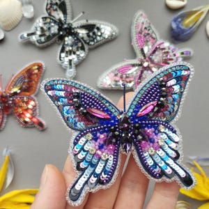 May include: A collection of handmade butterfly brooches. The brooches feature intricate beadwork and sequin details in various colors, including blue, purple, pink, and orange. The butterflies are displayed on a gray surface.