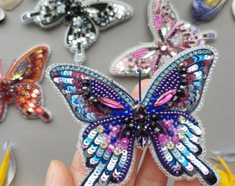 Può includere: Una collezione di spille a forma di farfalla fatte a mano. Le spille presentano intricati dettagli di perline e paillettes in vari colori, tra cui blu, viola, rosa e arancione. Le farfalle sono esposte su una superficie grigia.