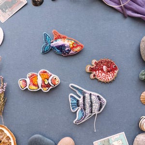 May include: Four colourful beaded fish brooches are displayed on a blue-grey surface. The brooches include a clownfish, a pufferfish, and two other fish designs with detailed beadwork. The brooches are embellished with sequins and beads.