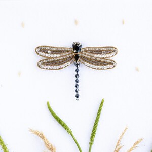 a decorative dragonfly brooch with a black body and white wings, surrounded by green leaves and small grains of rice against a white background.