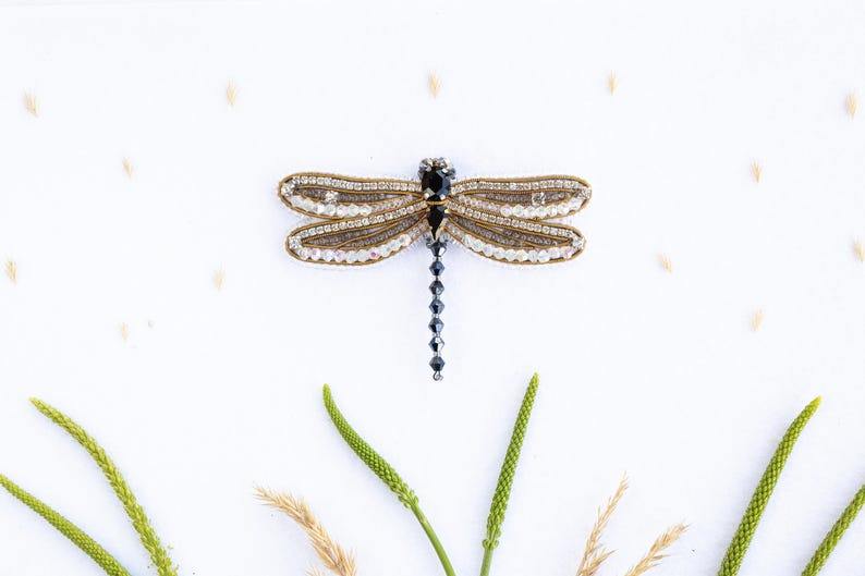 a decorative dragonfly brooch with a black body and white wings, surrounded by green leaves and small grains of rice against a white background.