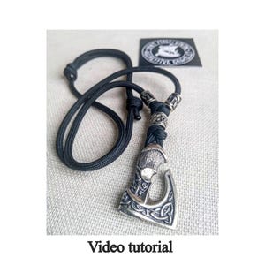 May include: A silver axe pendant with intricate knotwork design, hanging from a black paracord necklace. The pendant is attached to the necklace with silver beads.