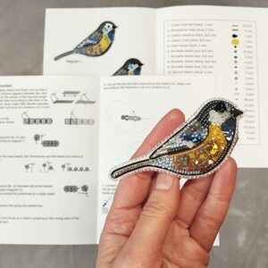 A hand holding a small, colorful bird-shaped embroidery pin or brooch, with instructions or diagrams visible in the background.