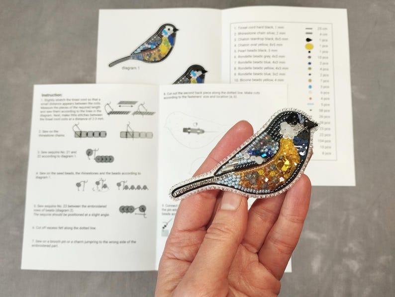 A hand holding a small, colorful bird-shaped embroidery pin or brooch, with instructions or diagrams visible in the background.