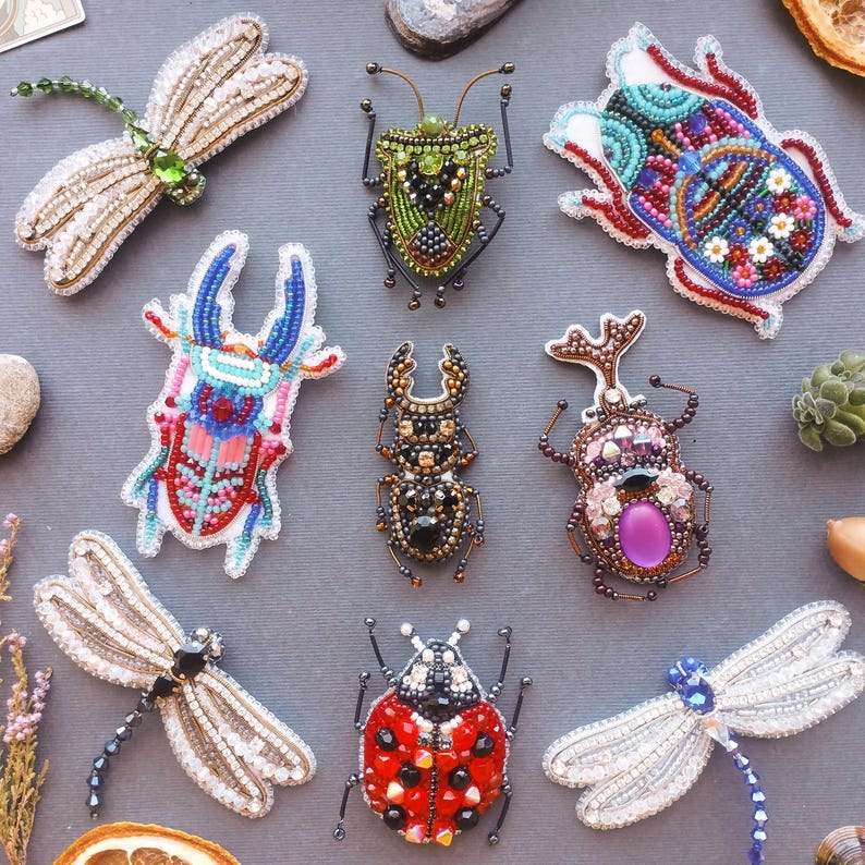 a collection of nine intricately designed, colorful, and detailed insect brooches or badges, each with unique patterns and embellishments.