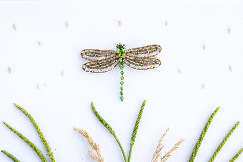 a decorative dragonfly brooch with green gemstones, surrounded by various natural elements such as grass, leaves, and small grains of sand.