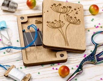 May include: A wooden sewing box with a hinged lid featuring a floral design. The box has a magnetic closure and contains a compartment with a blue thread and several needles. The box is sitting on a white wooden surface.