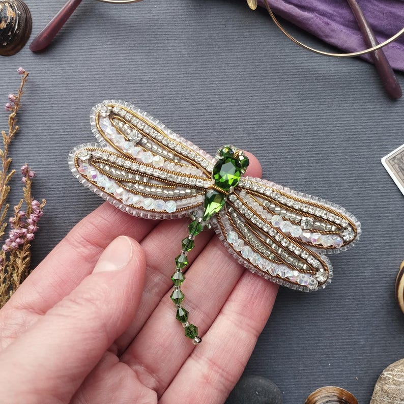 A hand holding a decorative brooch featuring a green gemstone and intricate beading.