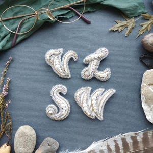 Peut inclure: Quatre broches de lettres perlées faites à la main : V, Z, S et W. Chaque lettre est blanche avec des perles en forme de perles, des accents dorés et une bordure perlée argentée. Les broches sont présentées sur une surface gris foncé avec des éléments naturels.