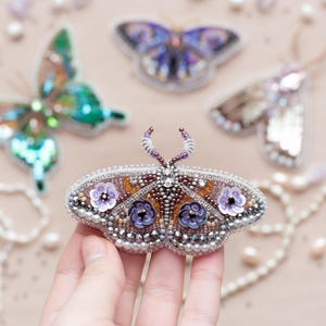 May include: Close-up of several beaded butterfly brooches in various colors, including green, purple, and brown. The brooches are embellished with sequins and beads, and are displayed on a light pink surface.