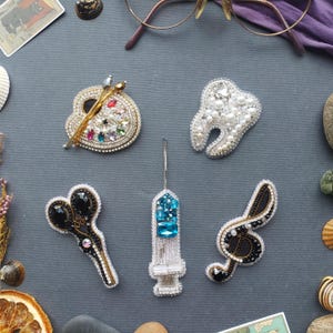May include: An assortment of handmade brooches, including a palette with paintbrushes, a tooth, scissors, a syringe, and a treble clef. Each brooch is intricately detailed with beads and crystals, set against a dark gray background.