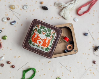 May include: A brown wooden box with a hinged lid. The lid is open and reveals a small, square, white fabric cushion with an embroidered design of two squirrels in a tree. The box is filled with sewing supplies, including spools of thread, needles, and beads.