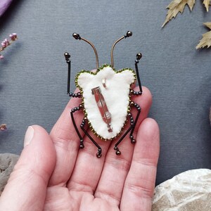 A hand holding a small, intricately designed insect-like object, which appears to be a beaded or embroidered piece.