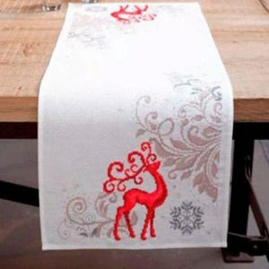 May include: White Christmas table runner with a red reindeer design. The runner features a detailed embroidered pattern of a reindeer, snowflakes, and floral accents. The runner is placed on a wooden table, adding a festive touch to any holiday decor.