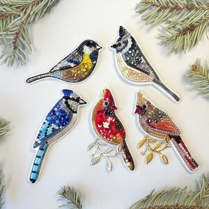 five colorful bird embroidered or beaded decorations arranged on a white background, surrounded by pine branches.