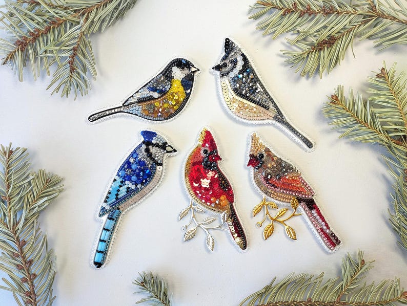 five colorful bird embroidered or beaded decorations arranged on a white background, surrounded by pine branches.