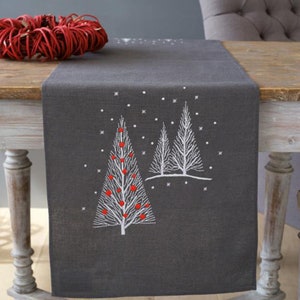 May include: A gray linen table runner with an embroidered design of two white trees with red ornaments. The trees are surrounded by white snowflakes.