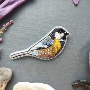 a decorative bird embroidered with colorful beads, surrounded by various natural elements such as leaves, flowers, and rocks.