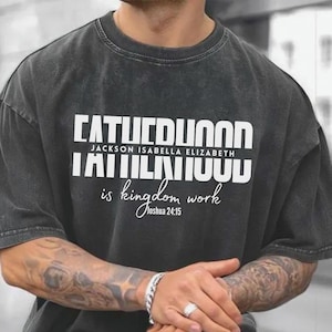May include: A dark gray t-shirt with the text "FATHERHOOD" in large white block letters. Below it, the text "JACKSON ISABELLA ELIZABETH" and "is kingdom work" in a script font. The shirt has a relaxed fit.